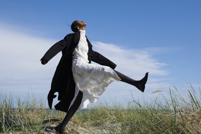a woman in a white dress and black coat jumping in the air