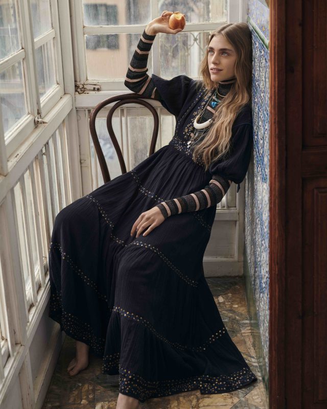 a woman in a long black dress leaning on a chair