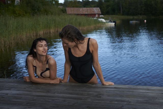 two women in swimsuits leaning on a dock by water
