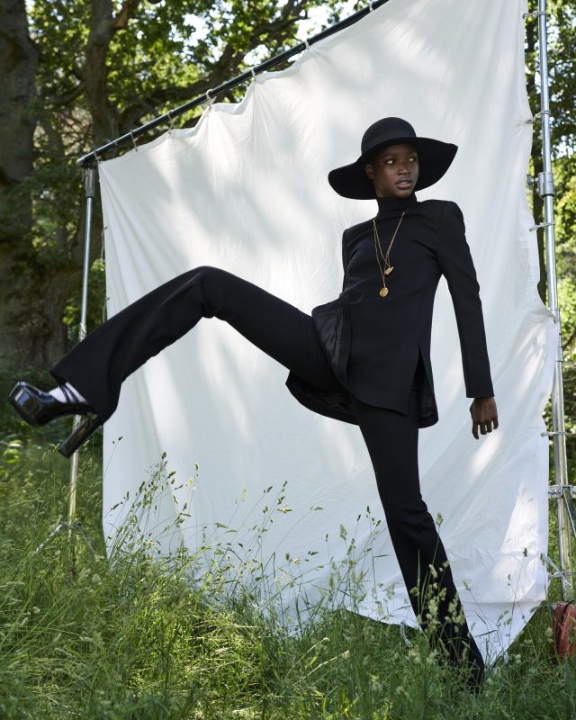 a woman in black outfit and hat standing in front of a white cloth