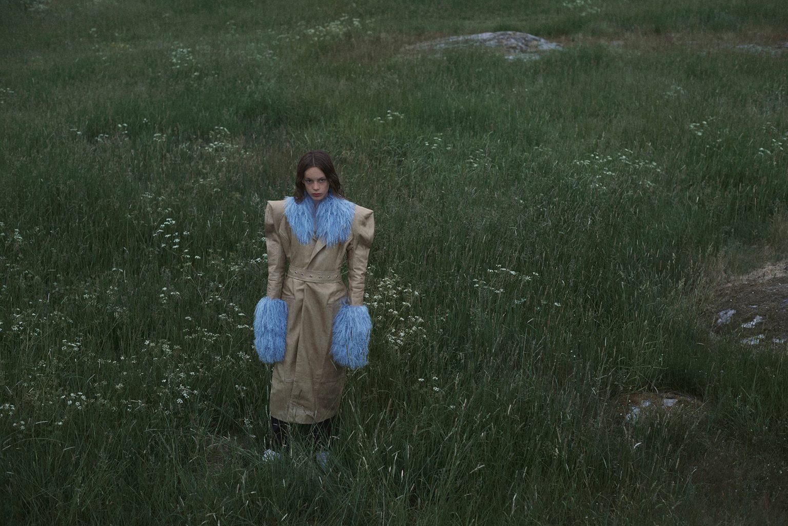 a woman in a long coat with blue furs standing in a grassy field