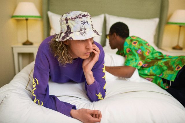 a man in a hat lying on a bed with another man in the background
