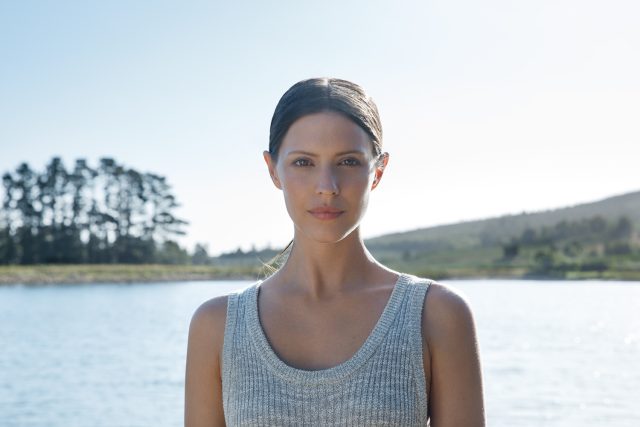 Woman in a gray tank top by a lake, looking at the camera.