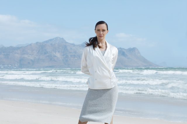 Woman in white jacket and grey skirt on a beach with mountains in background.