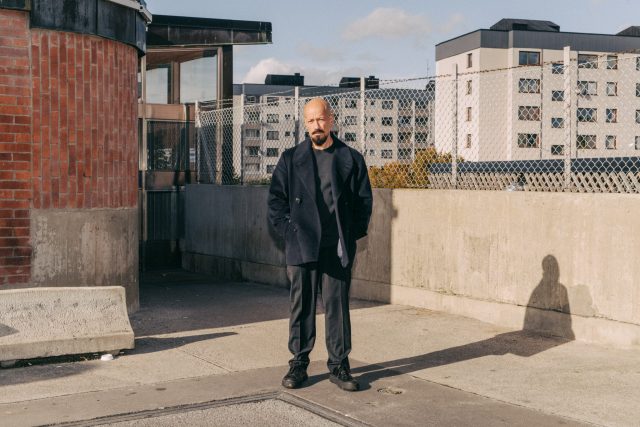 a man standing outside with a fence and buildings behind him