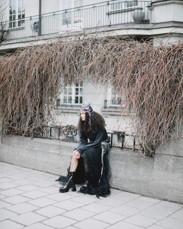 Woman in black dress and crown sits by a building with bare branches.