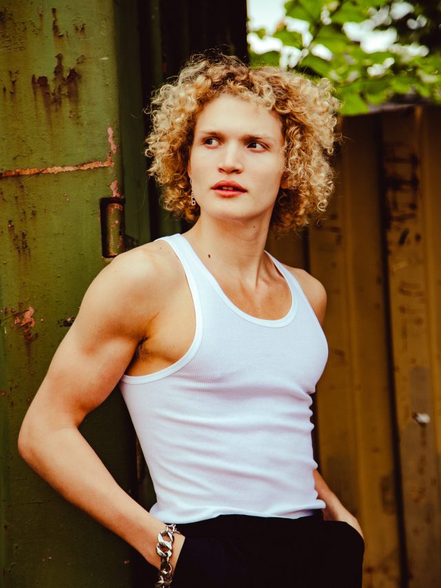 a man with curly blonde hair wearing a white tank top and blue jeans