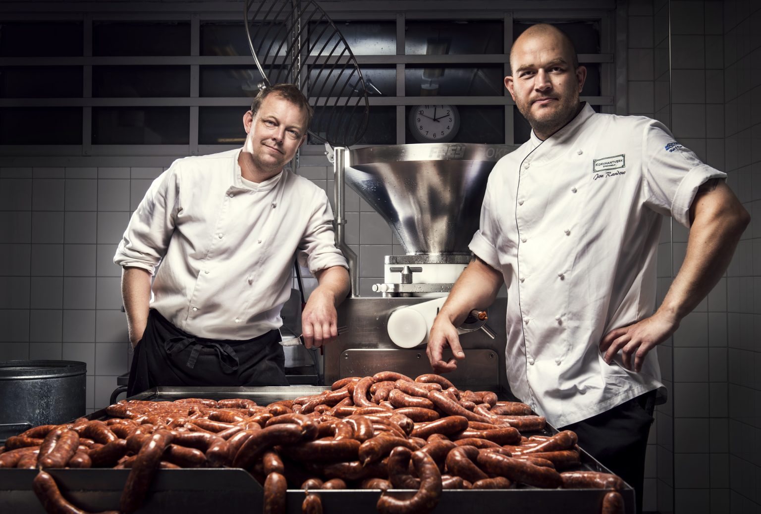 two men standing in a kitchen with sausages