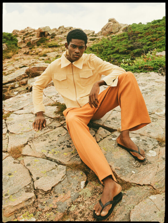 Young Black man in orange pants and yellow jacket sits on rocks.