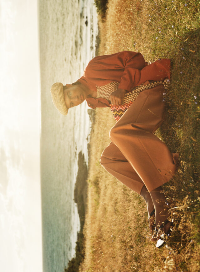 Person in red shirt and orange pants with a beaded bag, sitting by the sea.