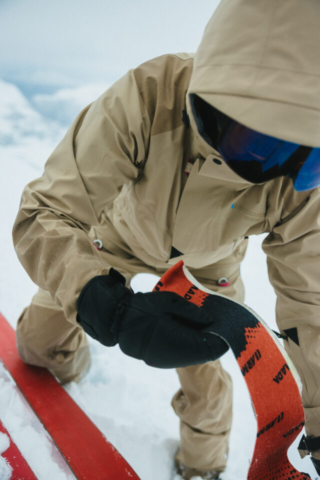 a person wearing a tan snow suit and goggles holding a black and red ski board