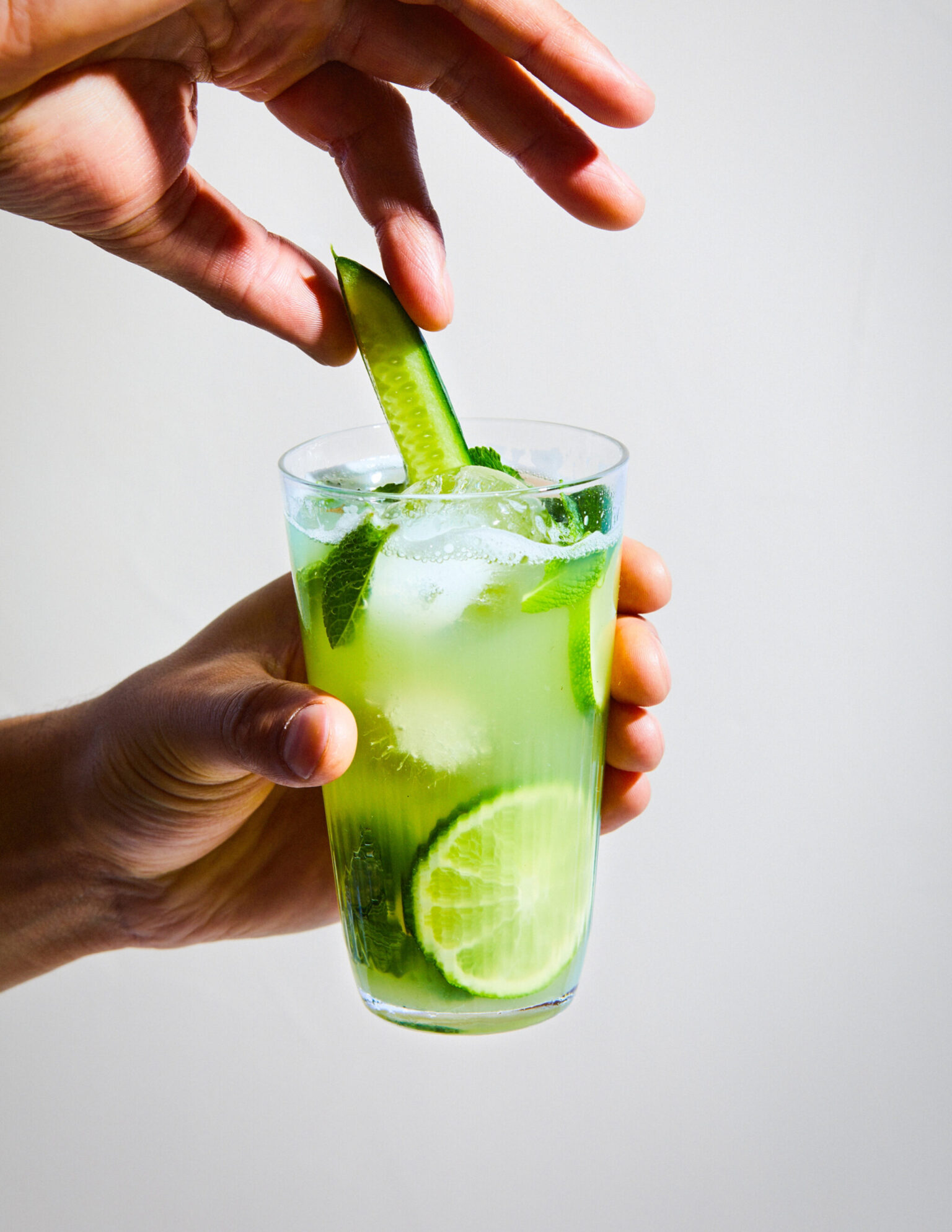 a person holding a glass of liquid with a cucumber and lime wedges