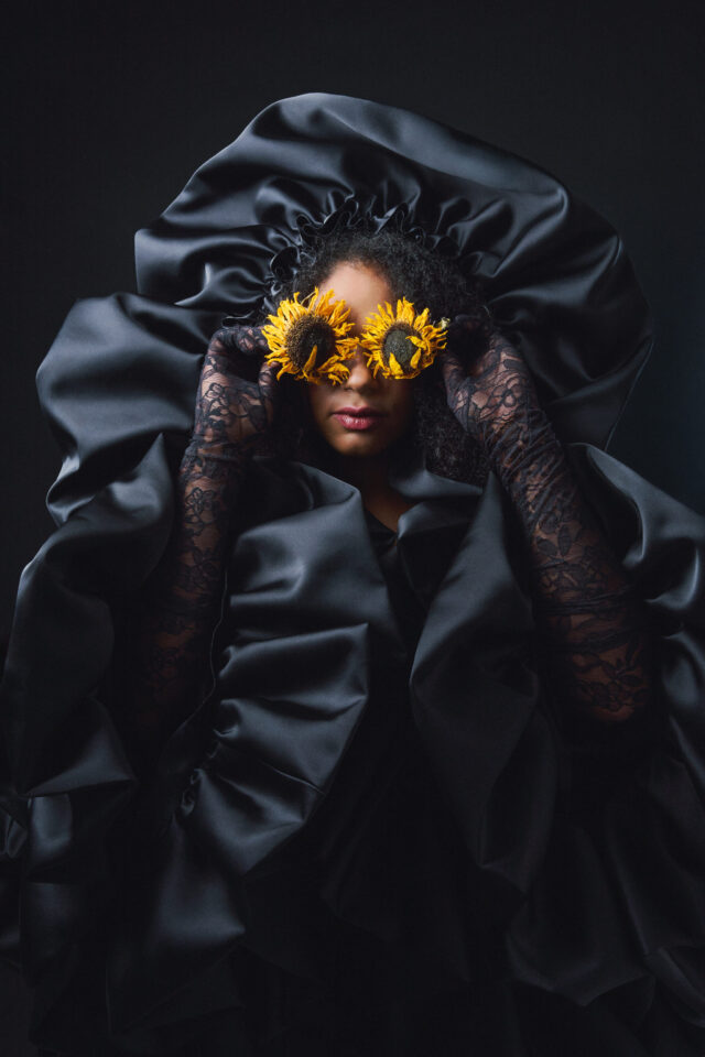 a woman wearing a black dress and holding sunflowers over her eyes
