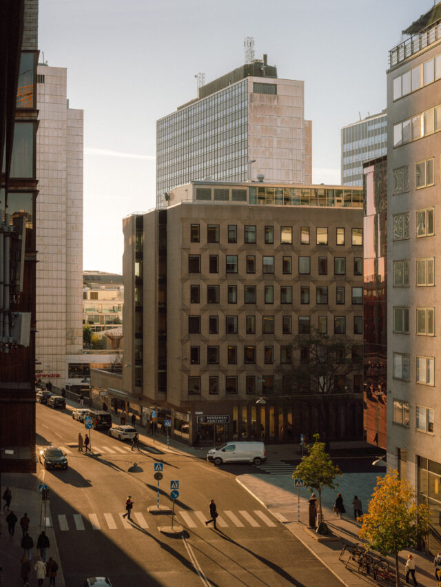 Street view of city buildings and people crossing a street at sunset.