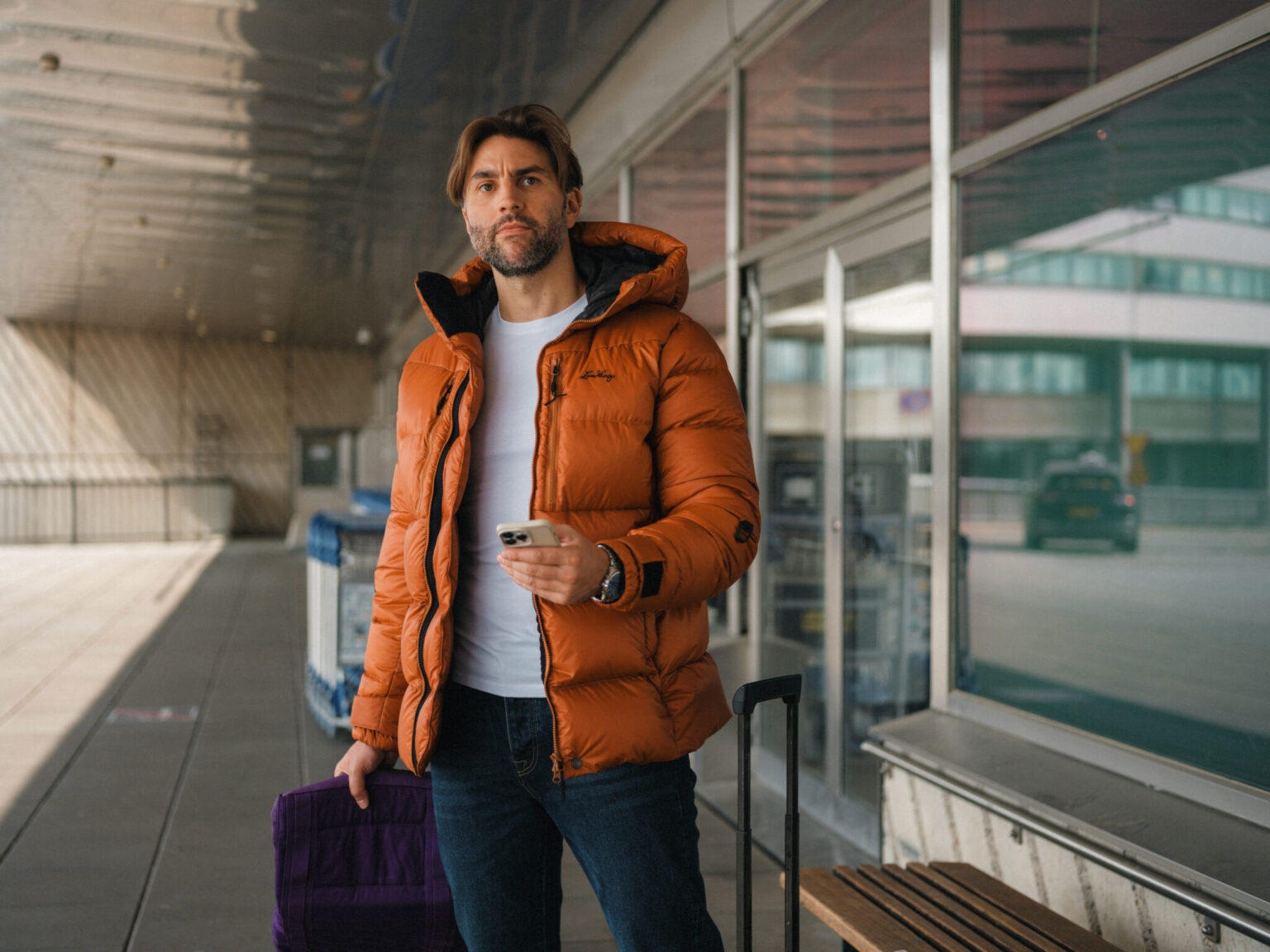Man in orange puffer jacket holding phone and purple bag at airport.