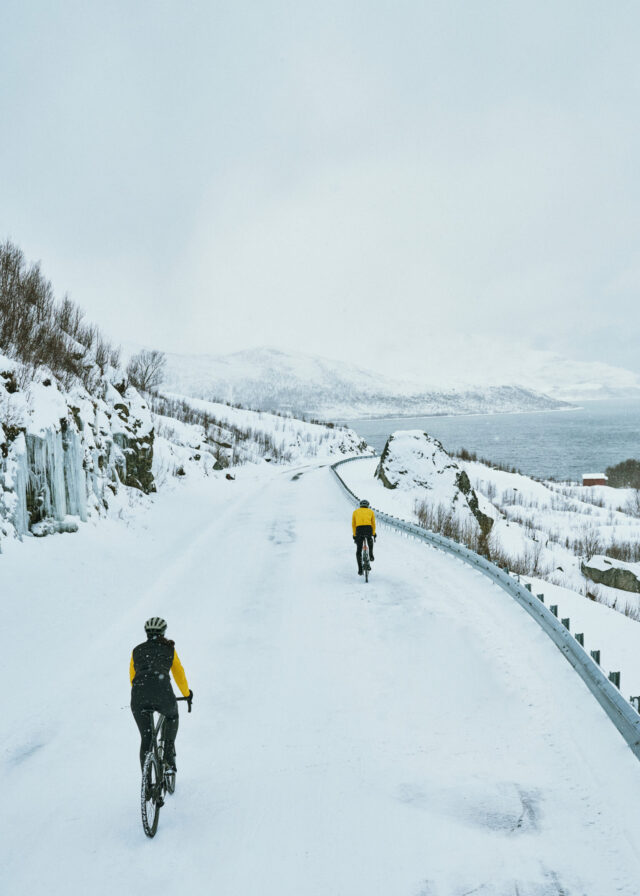 Two cyclists in yellow jackets ride on a snowy road by a lake.
