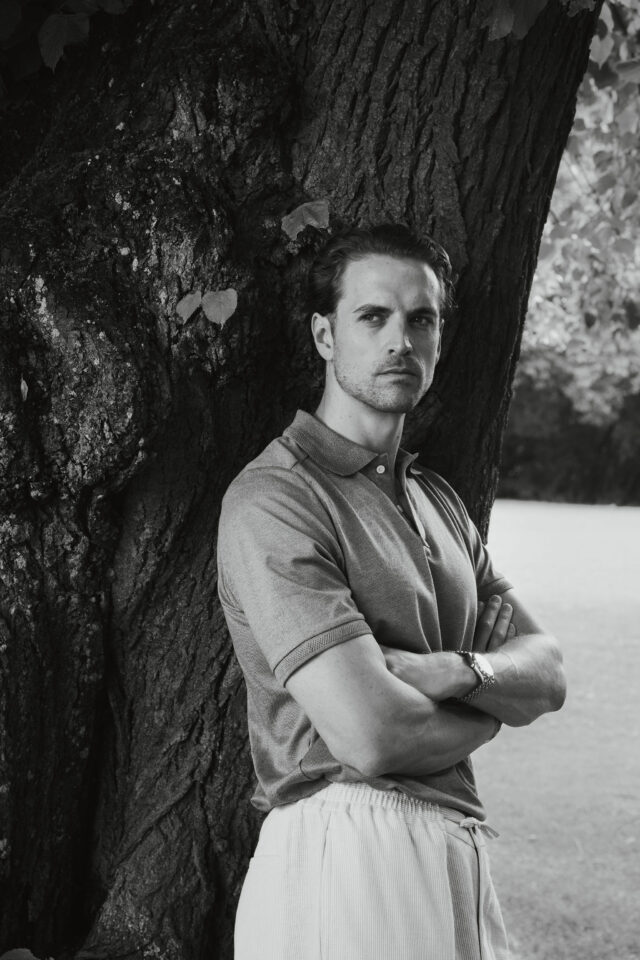 Man in polo shirt and striped pants leaning against a tree, arms crossed.