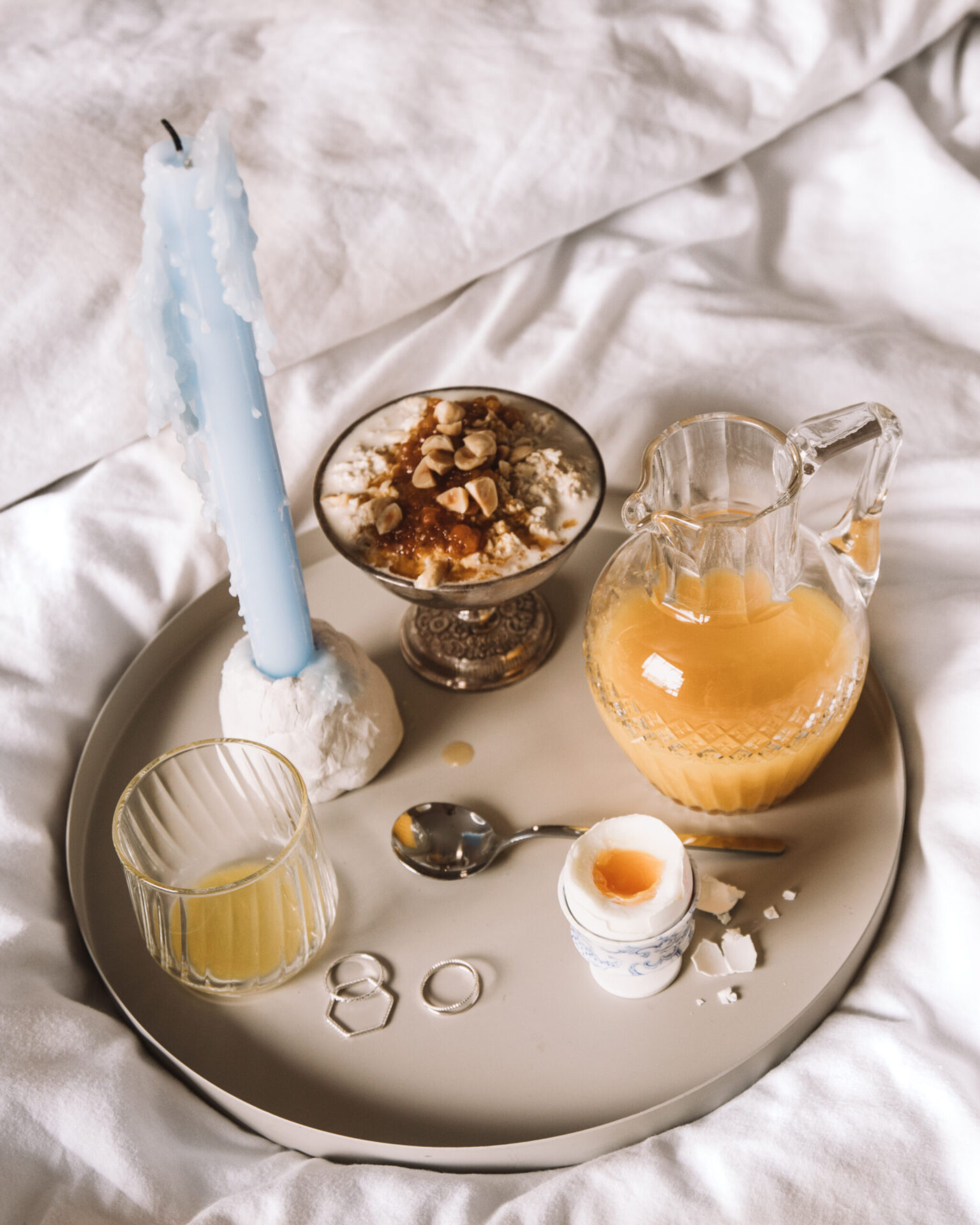 Breakfast tray with soft-boiled egg, juice, dessert, and a blue candle on a white linen background.