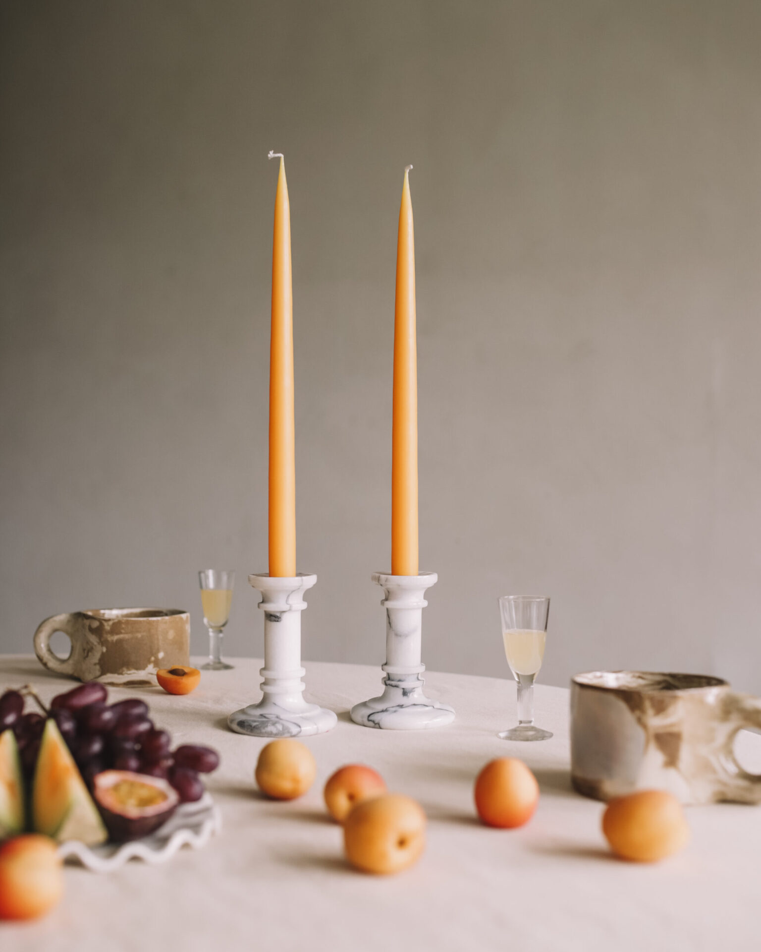 Two orange taper candles in marble holders on a table with fruit.