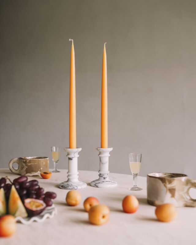 Two orange taper candles in marble holders on a table with fruit.