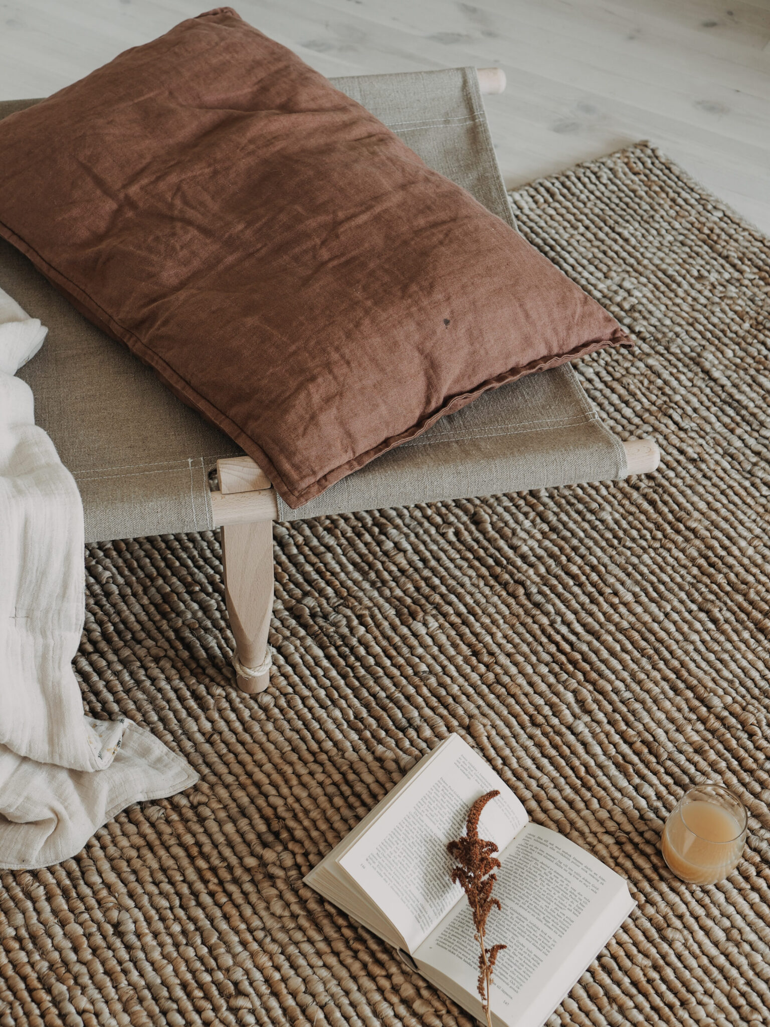 A brown pillow rests on a canvas chair over a woven rug, with a book and drink nearby.
