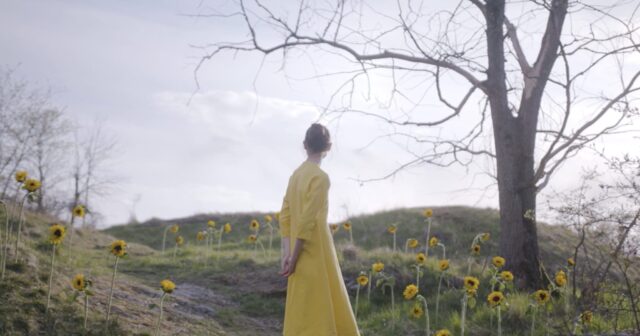 Woman in yellow dress in a field of sunflowers with bare trees.