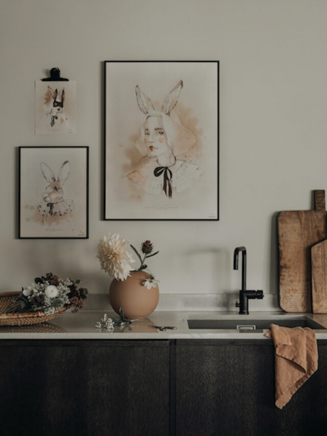 Framed watercolor art of a person with bunny ears hangs above a kitchen counter with flowers and a sink.