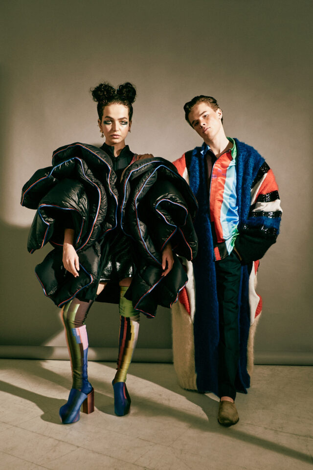 Two models in avant-garde fashion, one in a voluminous black puffer coat, the other in a colorful, textured coat.