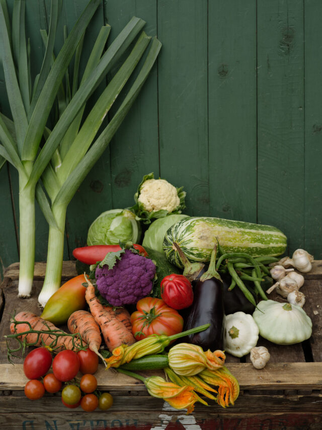 A rustic wooden crate filled with fresh, colorful vegetables including leeks, tomatoes, carrots, and squash.