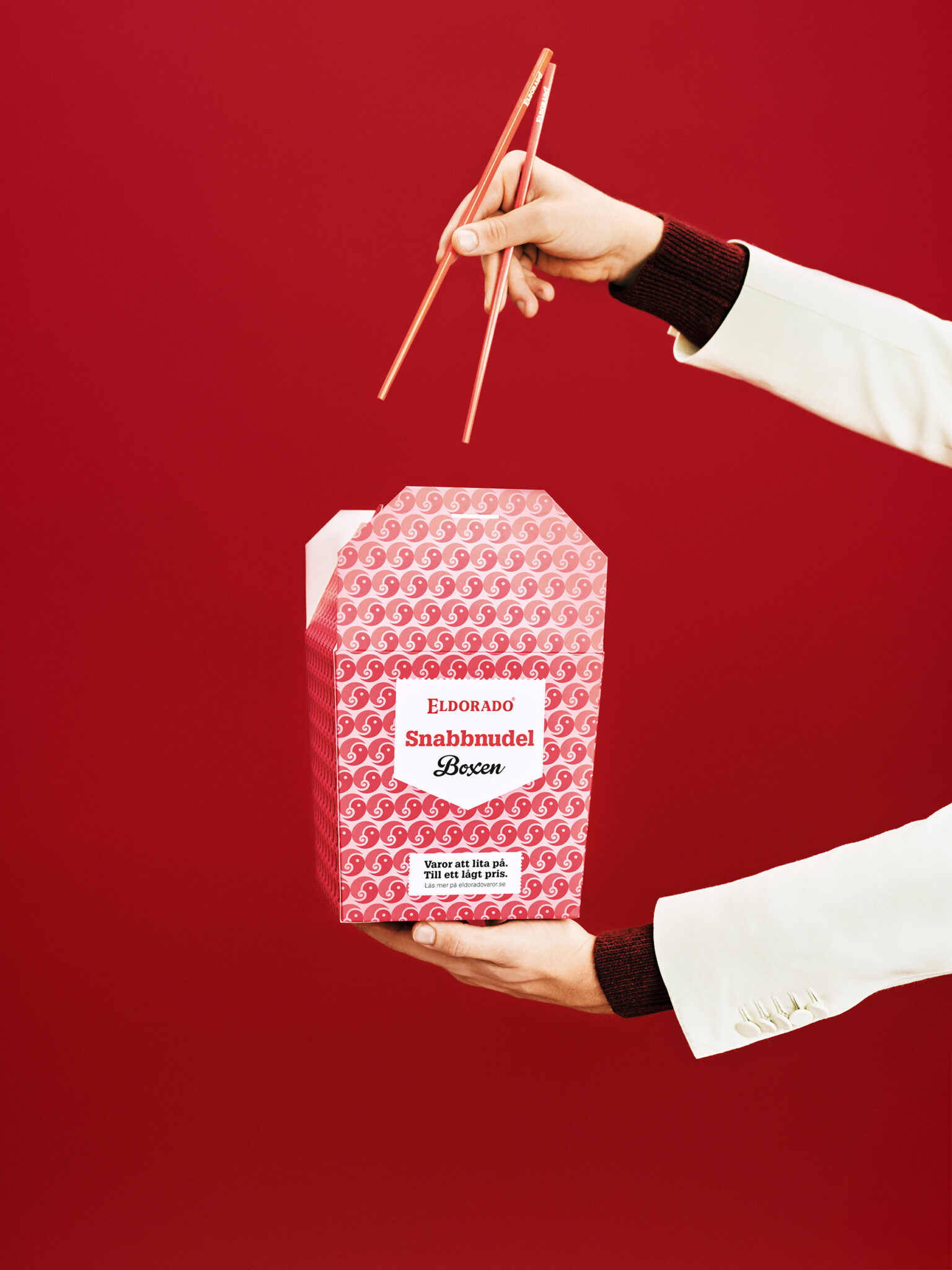 A hand holds a red and pink patterned noodle box with chopsticks above it.
