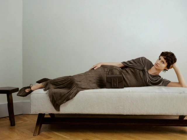 Woman in brown dress and shoes reclines on a white furry chaise lounge with a brown handbag.