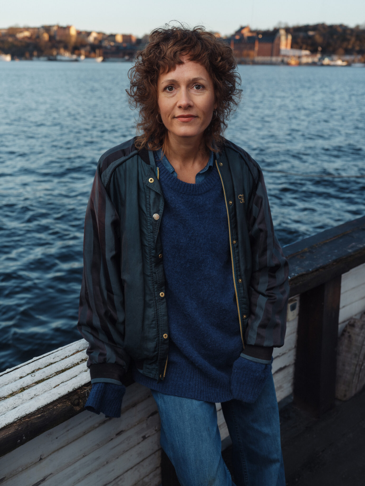 Woman with curly hair and a bomber jacket stands by the water.