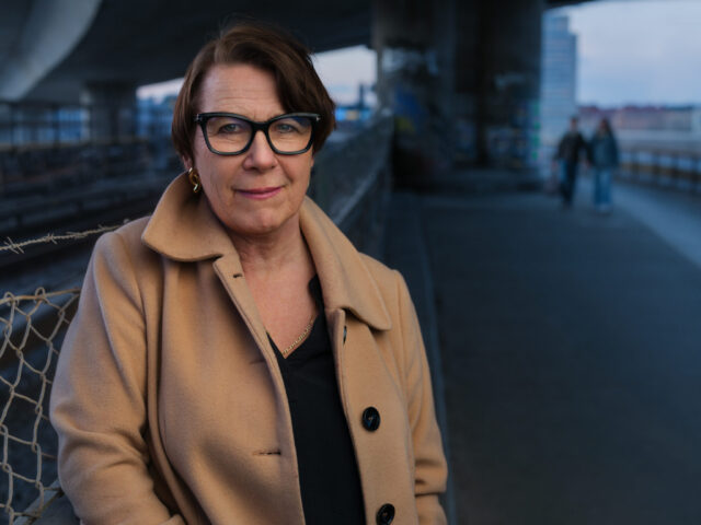Woman in a tan coat and glasses smiling.